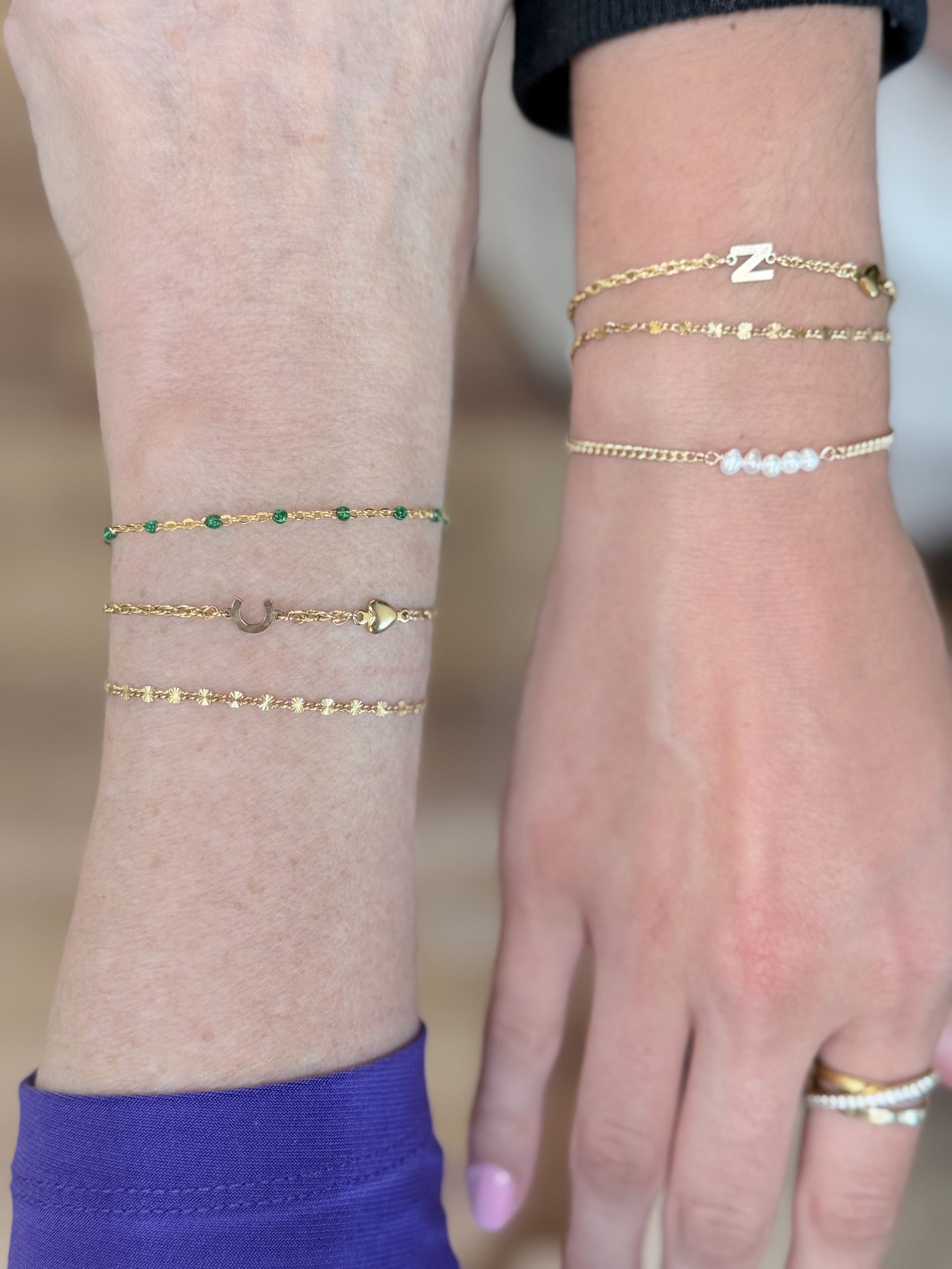 Two wrists layered with gold bracelets, a diamond letter Z charm, a seed-pearl bar bracelet, and a horseshoe-and-heart charm chain