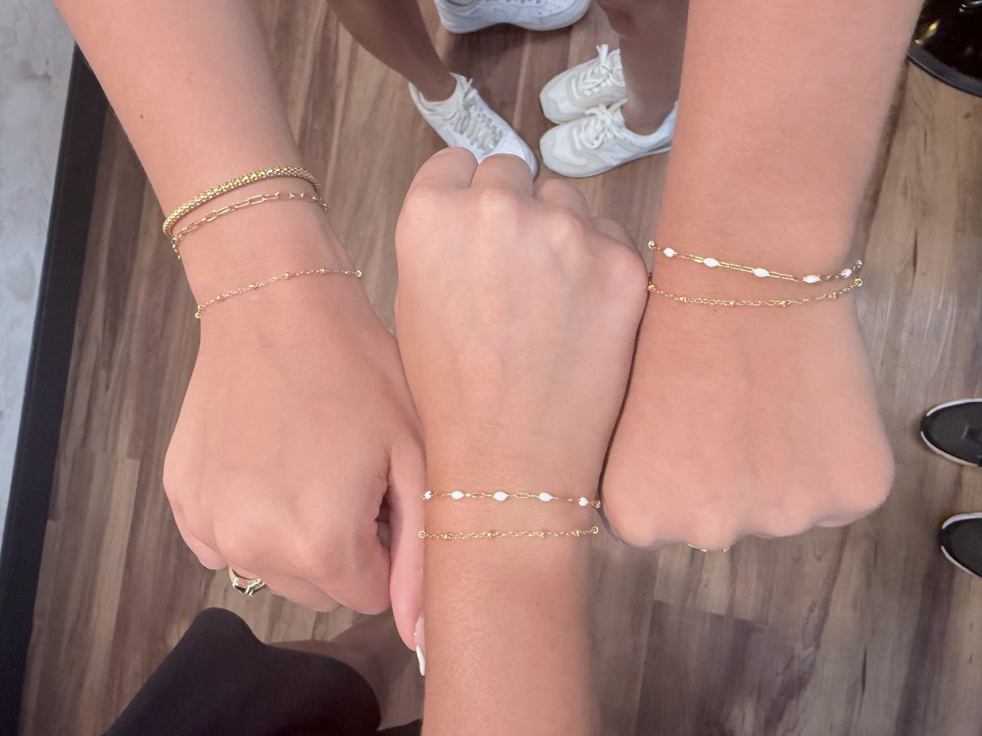 Three friends' fists stacked together, each wearing layered gold bracelets with textured bar links and white beaded chains