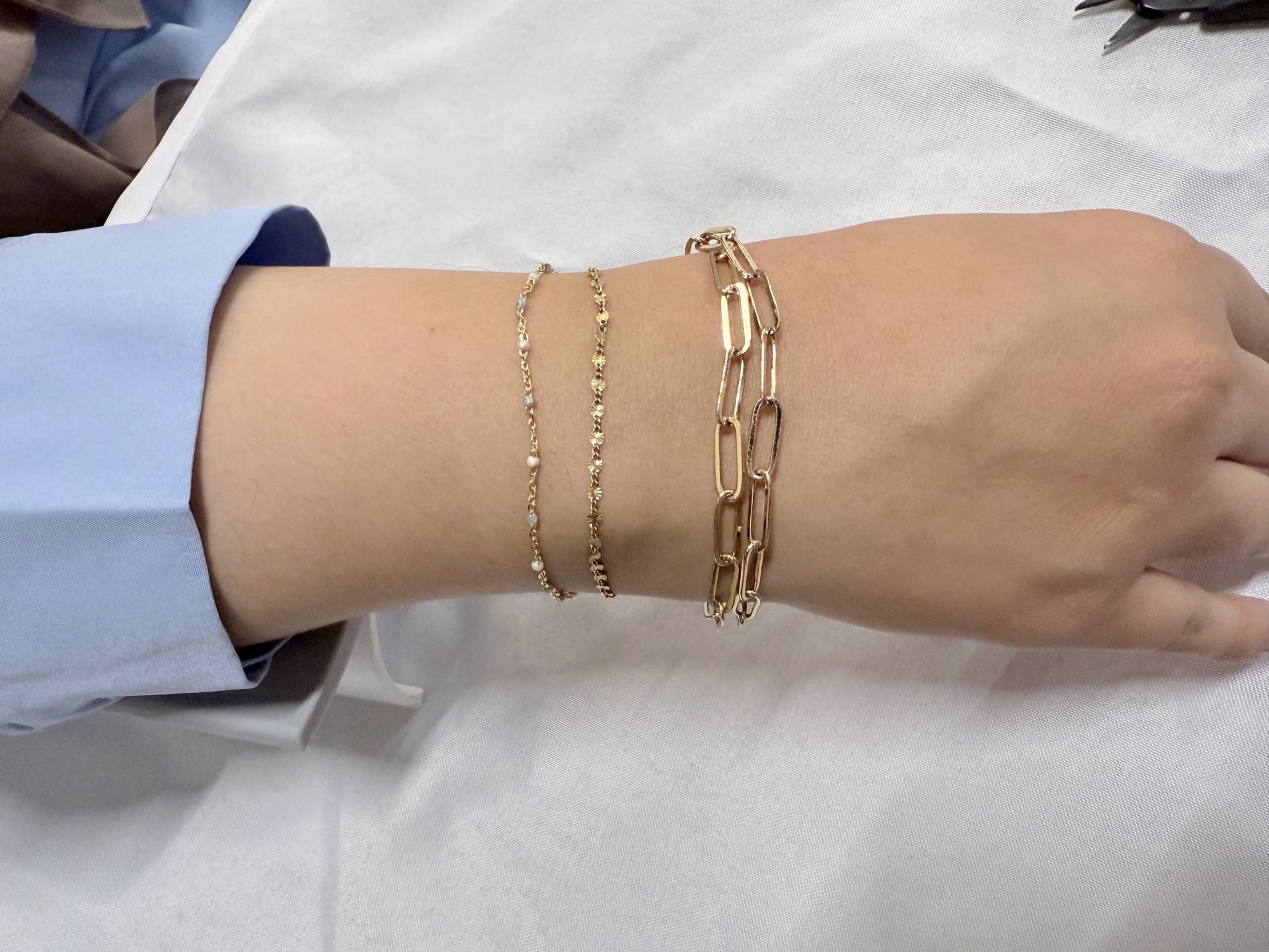 Wrist wearing three layered gold bracelets: a beaded chain, a textured curb chain, and a paperclip-link chain