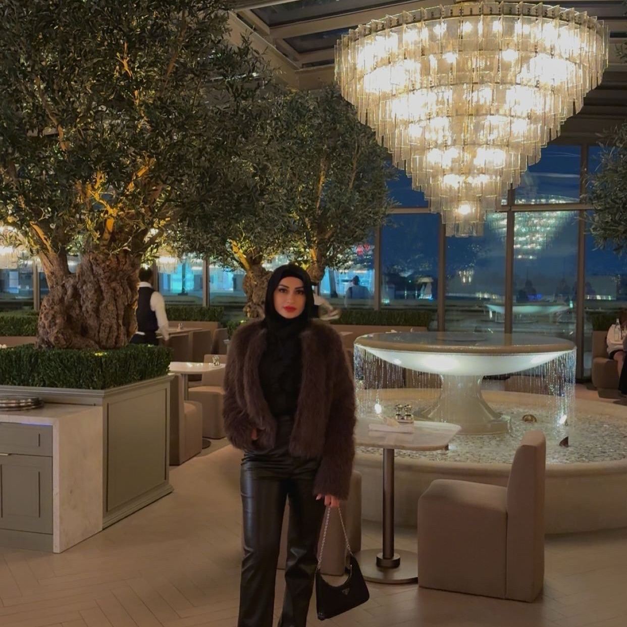 Huda dressed up at an evening event, surrounded by olive trees and a large crystal chandelier