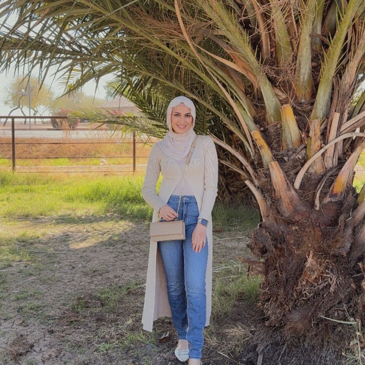 Huda outdoors in a casual sweater and jeans, smiling beside a palm tree