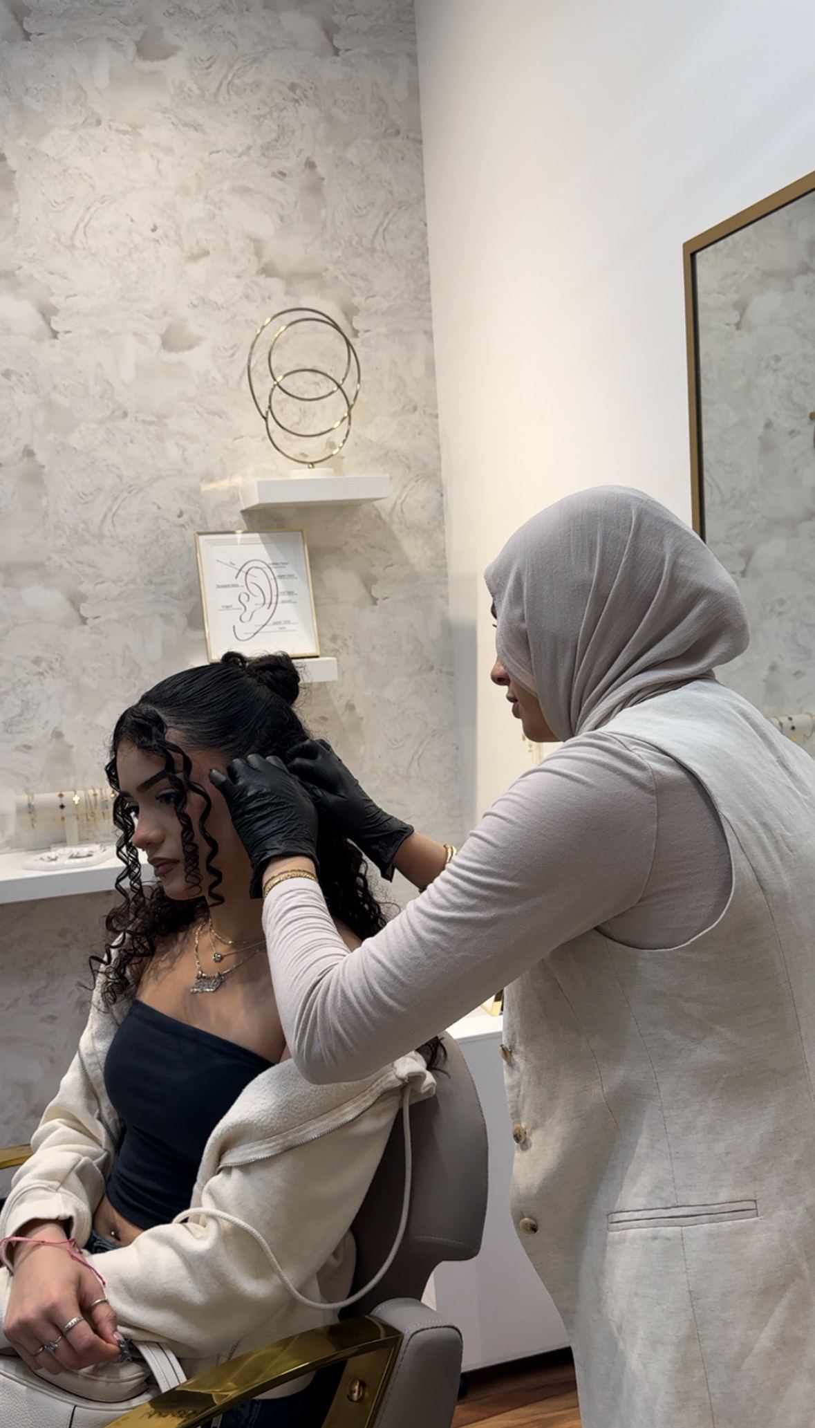 Huda piercing a seated client, studio shelves and a framed ear placement diagram visible behind her